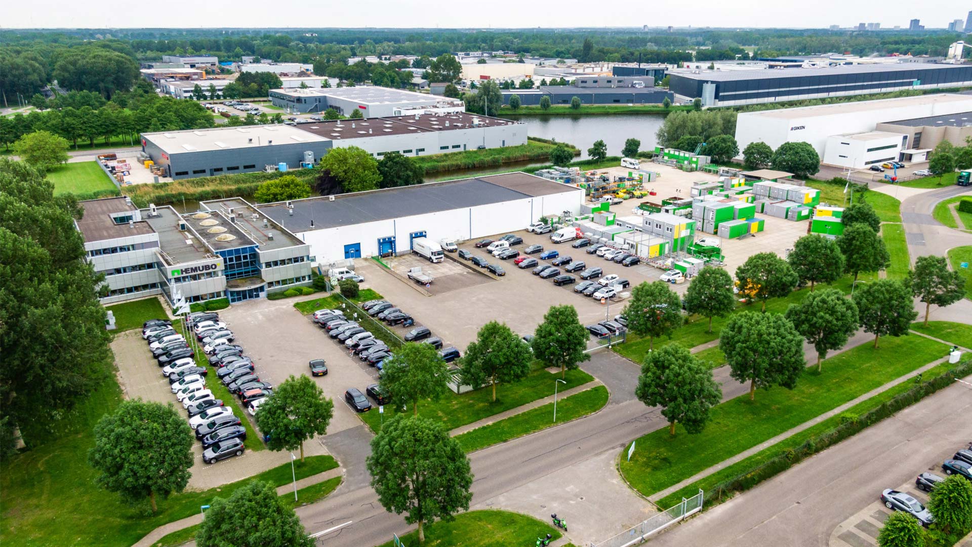 Luchtfoto van een bruisend industrieterrein met een georganiseerde indeling van gebouwen, parkeerplaatsen vol auto’s, groene stukken met bomen en omliggende wegen – een momentopname van stedelijke ontwikkeling en economische activiteit.