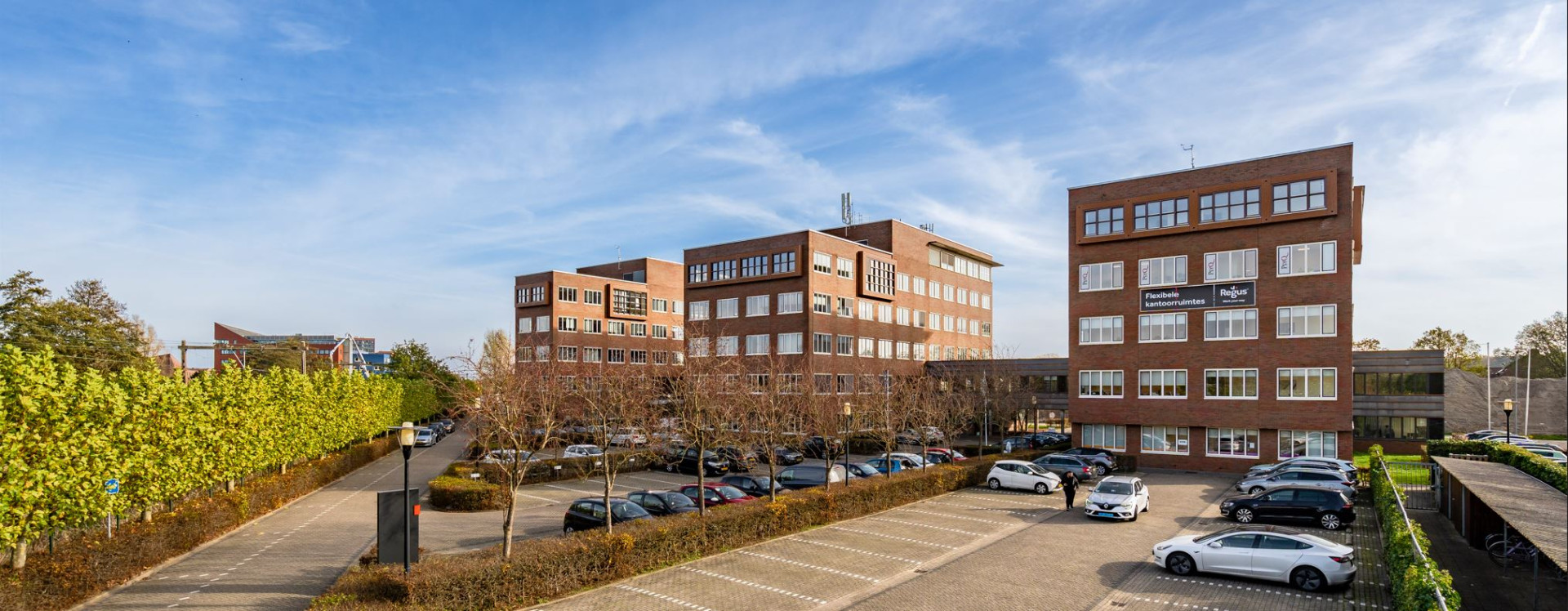 Een kantoorgebouw met een aan de gevel een bord met een aankondiging voor flexibele kantoorruimtes. Het gaat om een advertentie of promotie voor kantoorruimtes die flexibel kunnen worden gehuurd. "Regus" is een bedrijf dat wereldwijd flexibele werkplekoplossingen biedt, waaronder gedeelde kantoorruimtes, virtuele kantoren en vergaderruimtes.