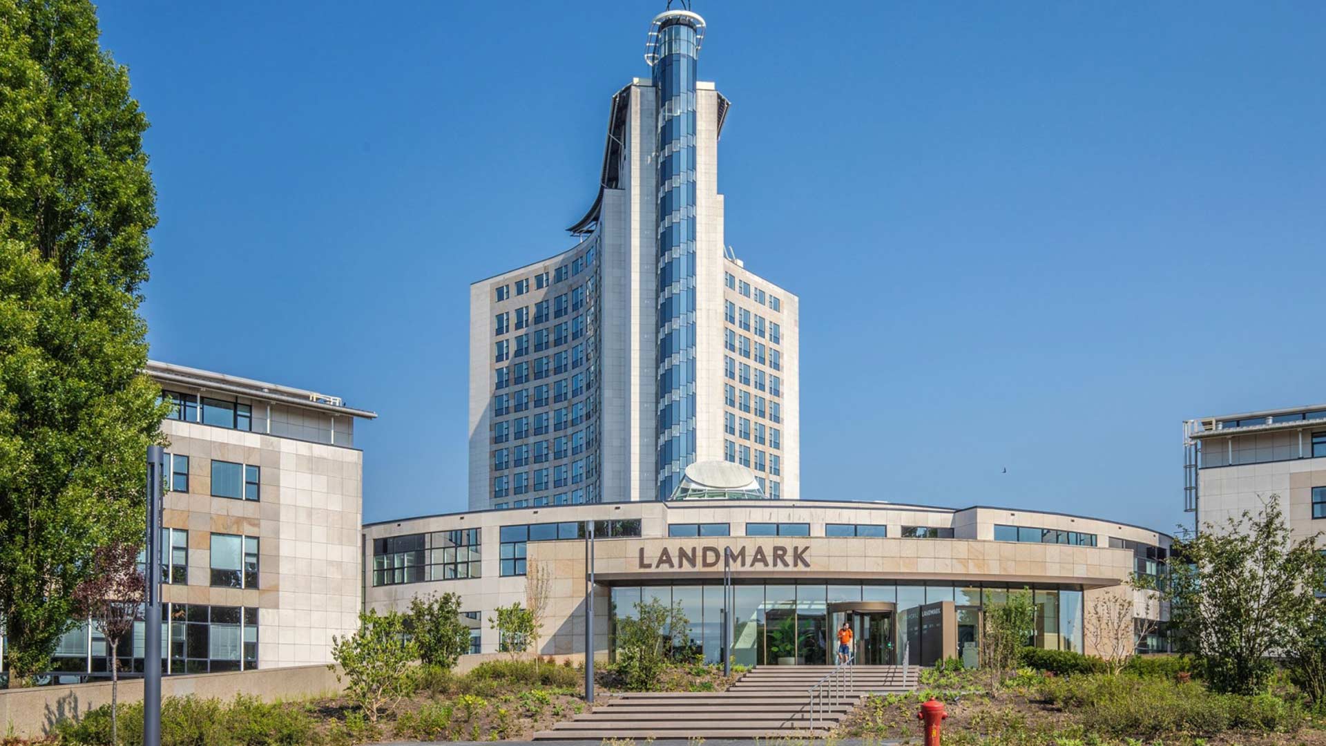 Een modern, glazen kantoorgebouw met een hoge toren en prominente spits, omgeven door bomen en groen, met de tekst 'LANDMARK' op de voorkant.