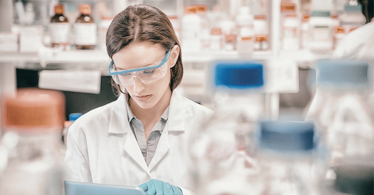 A scientist in a lab coat and safety glasses carefully reviews data on a tablet computer.