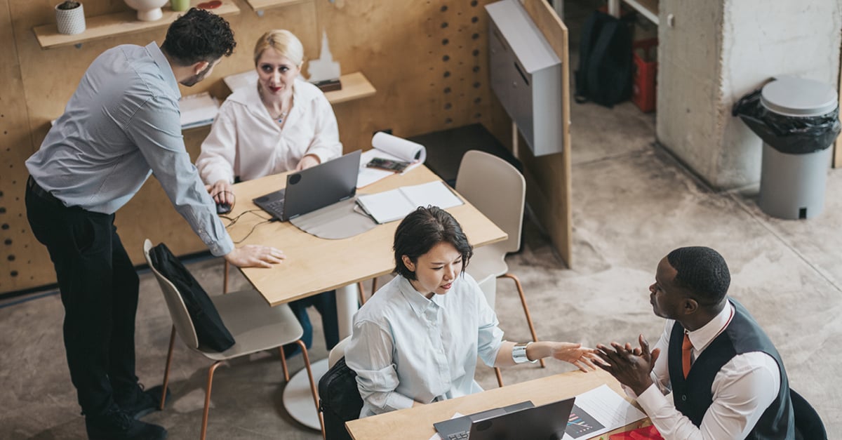 Overhead shot of a diverse team of four colleagues actively working and discussing ideas at adjacent desks with laptops and documents in a creative open-plan commercial office space.