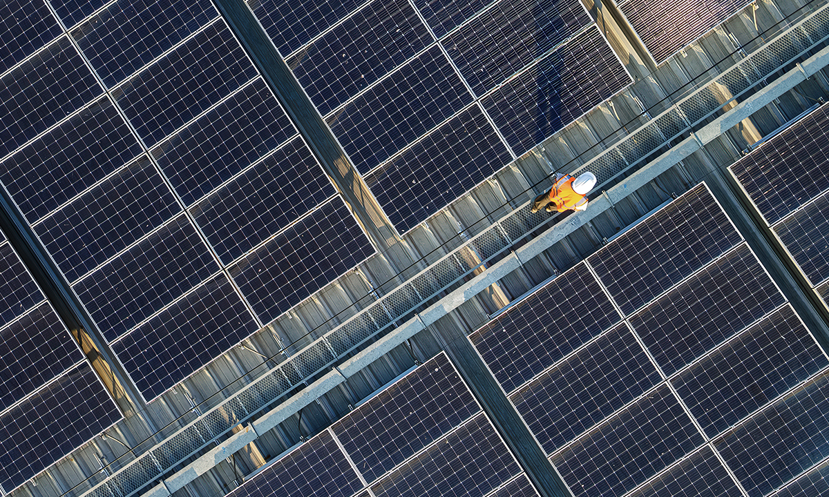 Aerial view of rooftop solar panels arranged in rows, with a worker in high‑visibility safety gear walking along a maintenance pathway between the panels.