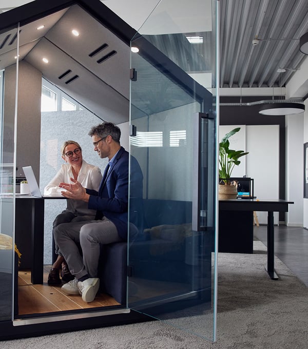 Two colleagues sitting inside a glass meeting pod in a modern office, discussing work while using a laptop.