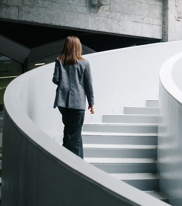 Person walking up a modern curved staircase inside a contemporary office building with concrete and metal architectural elements.