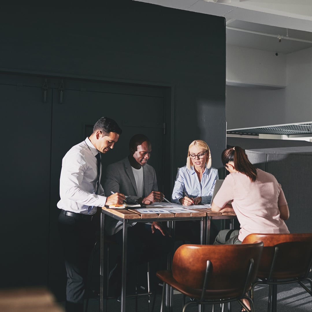 Group of people collaborating at a high table in a modern office environment with documents spread out for discussion.