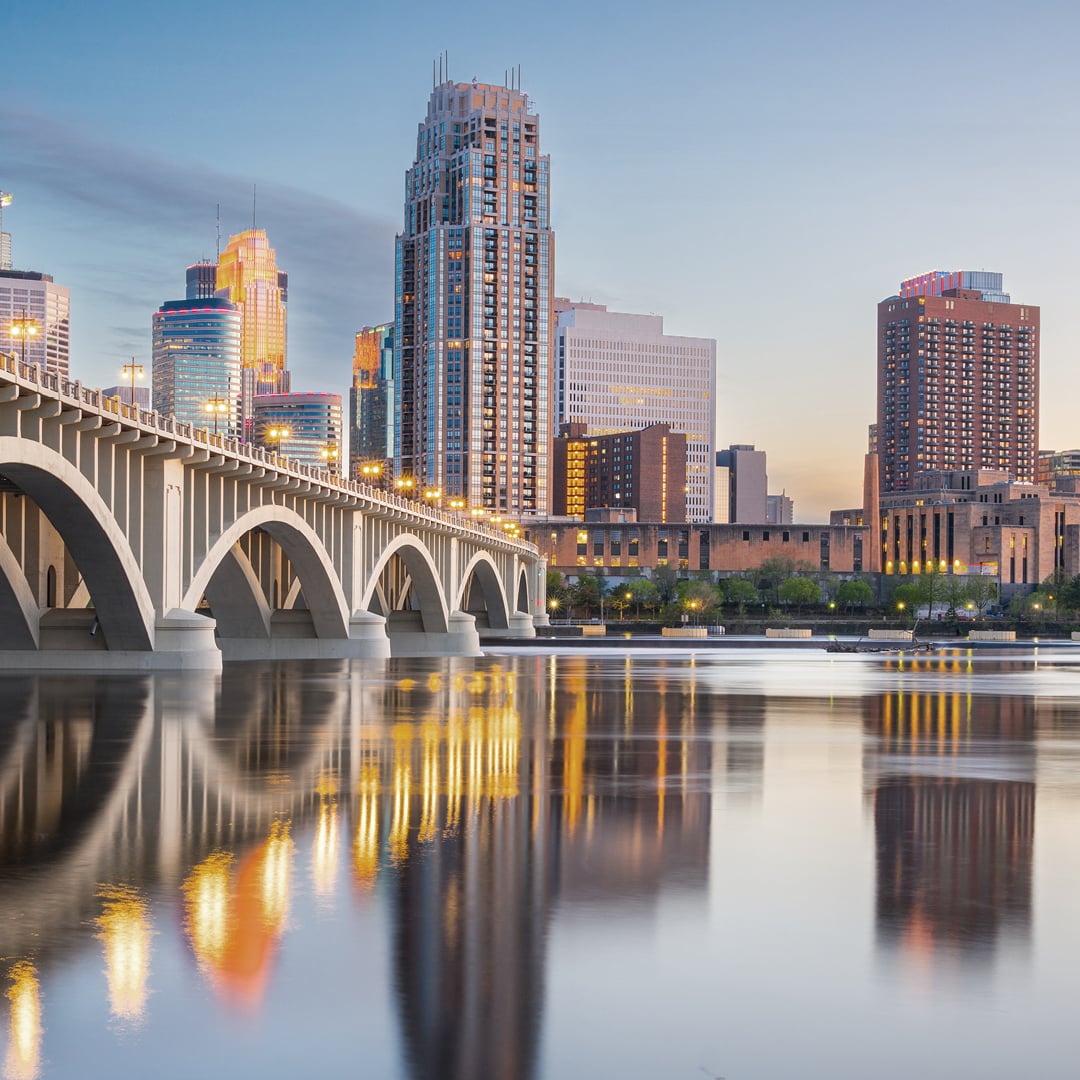 A beautiful dusk view of the Minneapolis skyline, featuring the illuminated Stone Arch Bridge spanning the Mississippi River with city lights reflecting on the water.