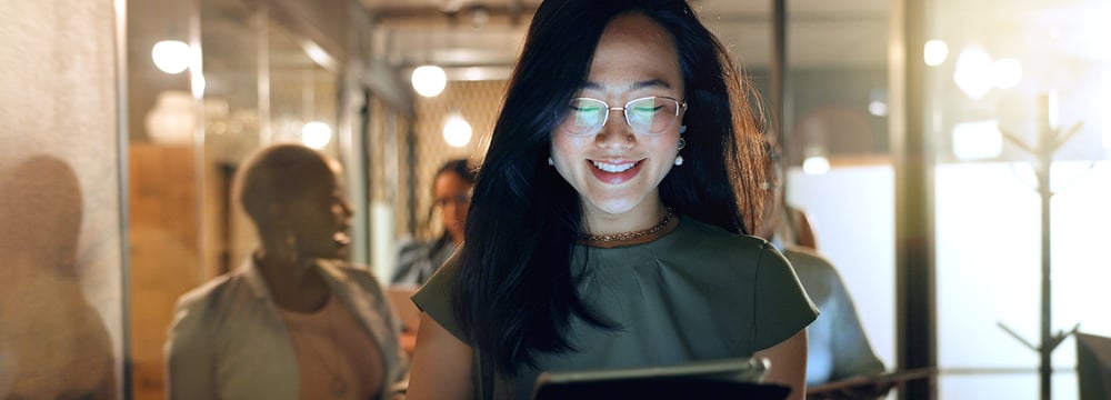 A smiling professional woman wearing glasses reviews data on a tablet while walking through a modern, warmly lit office corridor with colleagues in the background.