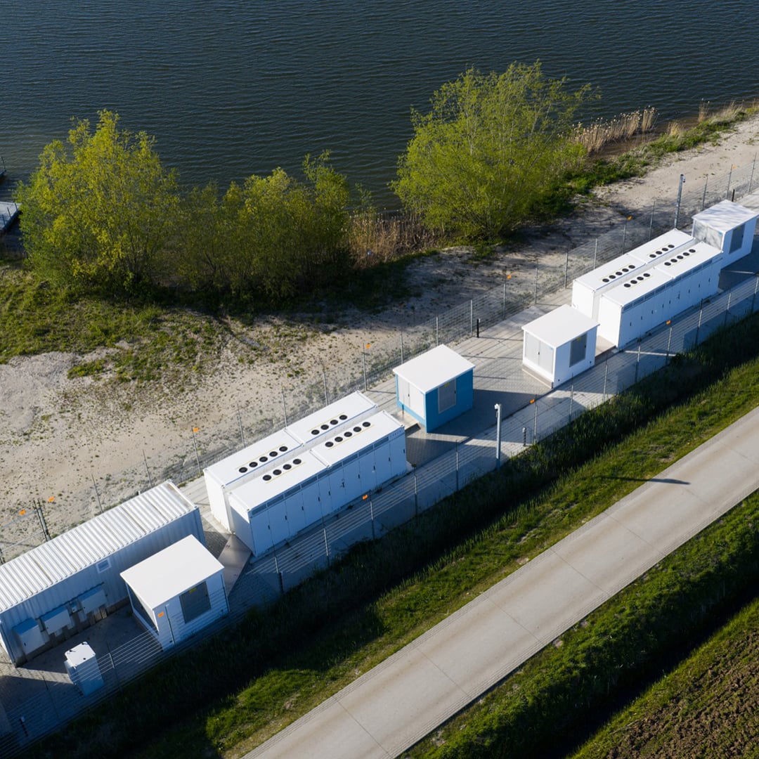 Aerial view of a distributed generation battery energy storage system with rows of white containerized BESS units in a fenced waterfront compound.