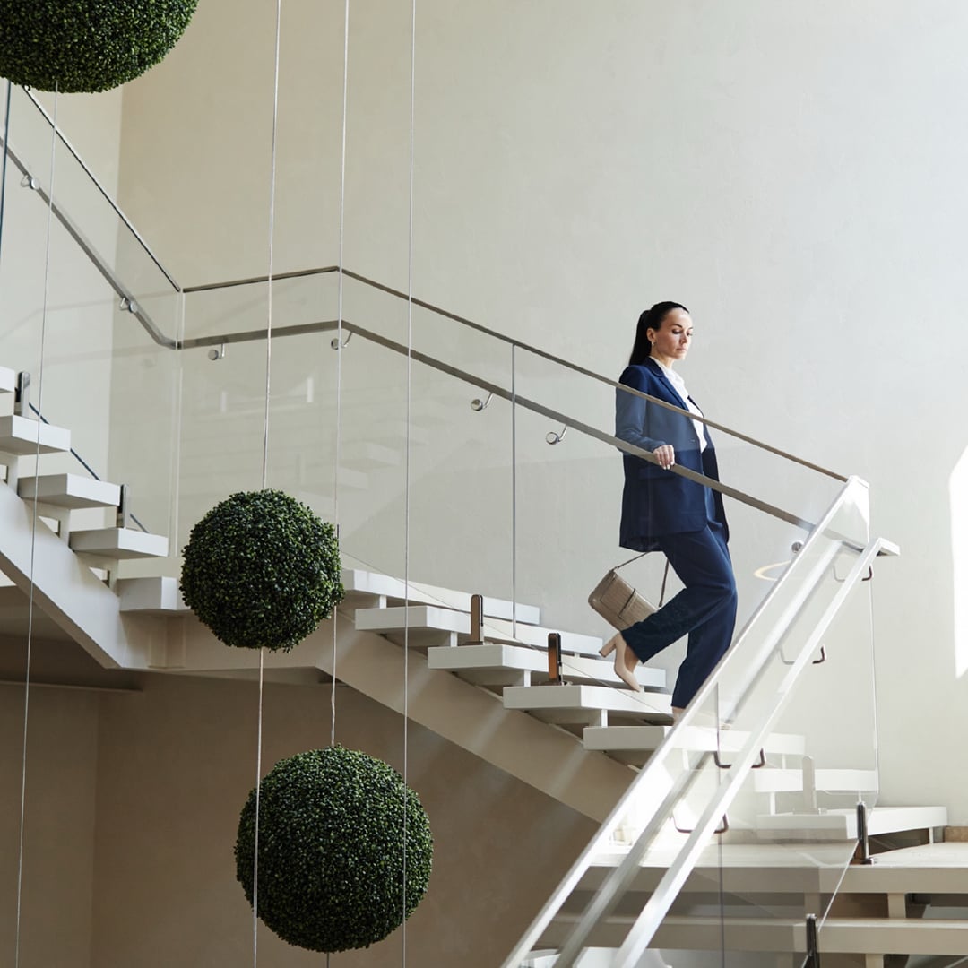 A professional woman in a blue suit ascends a modern, elegant staircase with white steps and glass railings in a stylish commercial building.