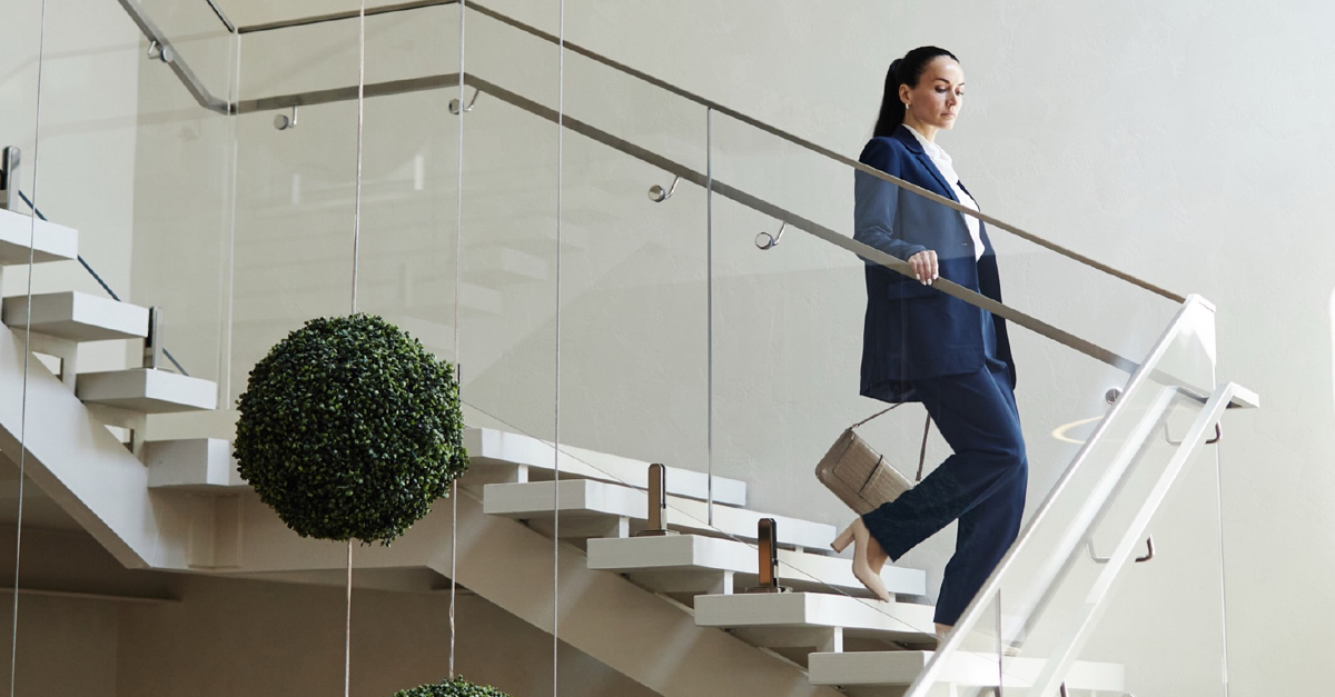 A professional woman in a blue suit ascends a modern, elegant staircase with white steps and glass railings in a stylish commercial building.
