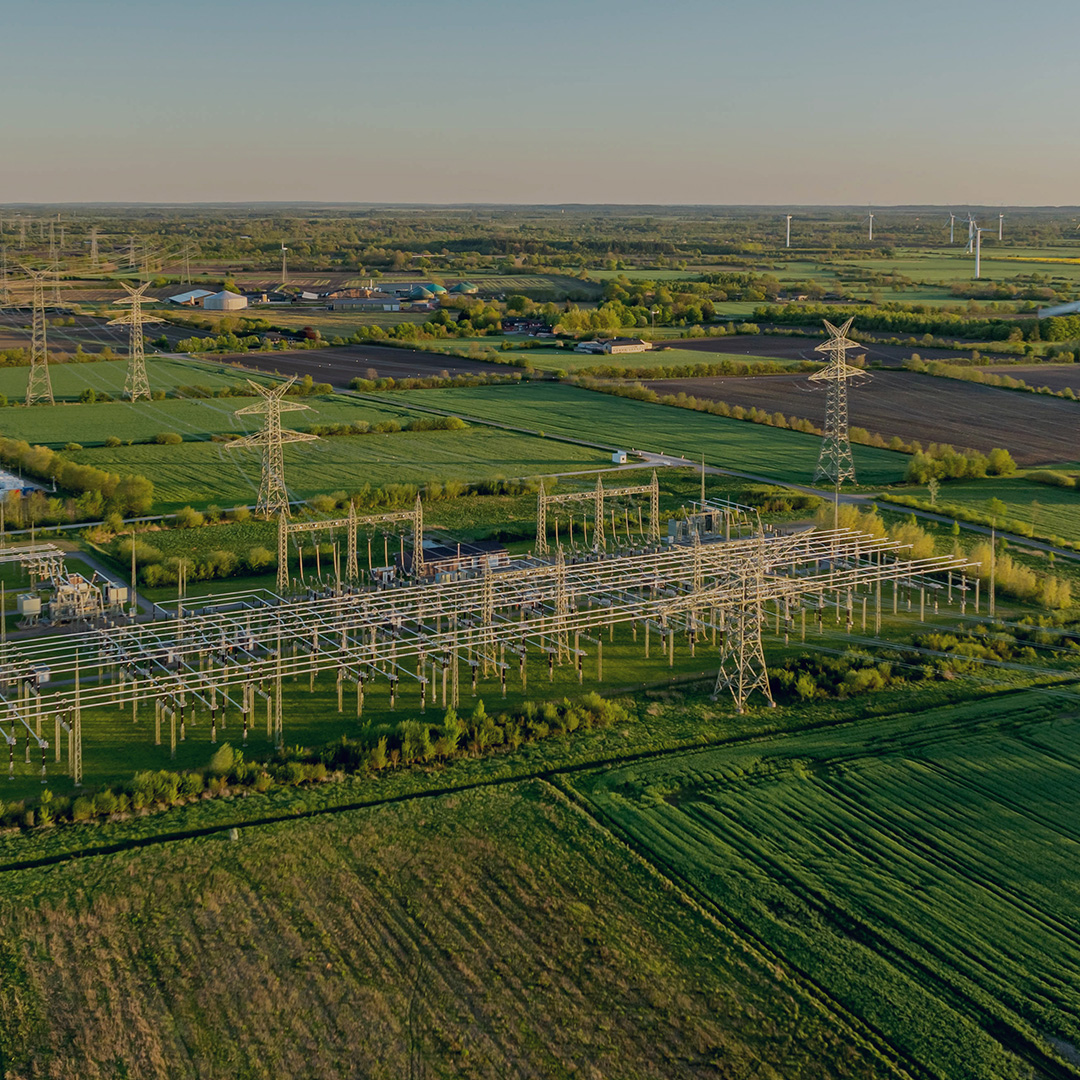 Aerial view of a rural electrical power substation with battery energy storage units and wind turbines across green agricultural fields at golden hour.