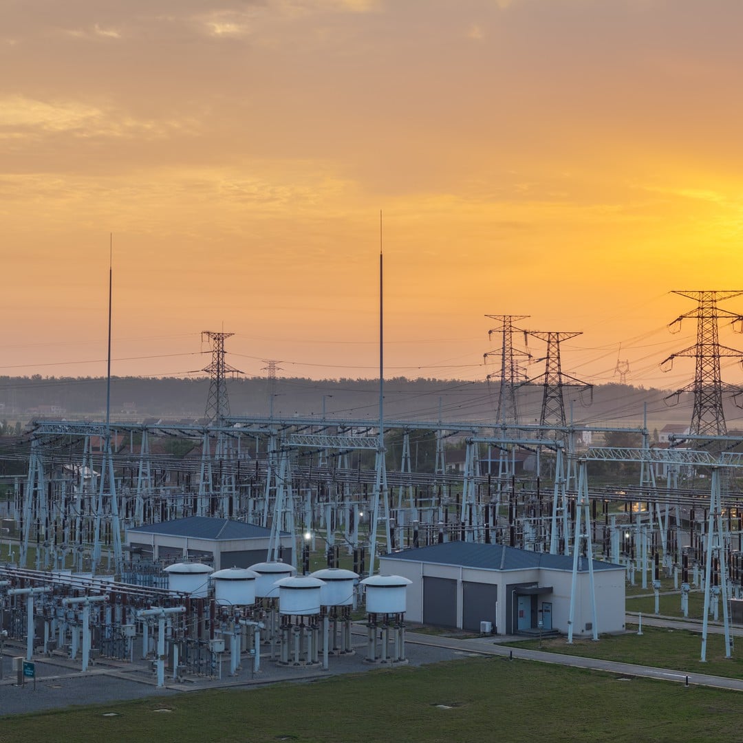 Aerial view of a large electrical substation with transformers and steel transmission towers silhouetted against a warm golden sunset sky.