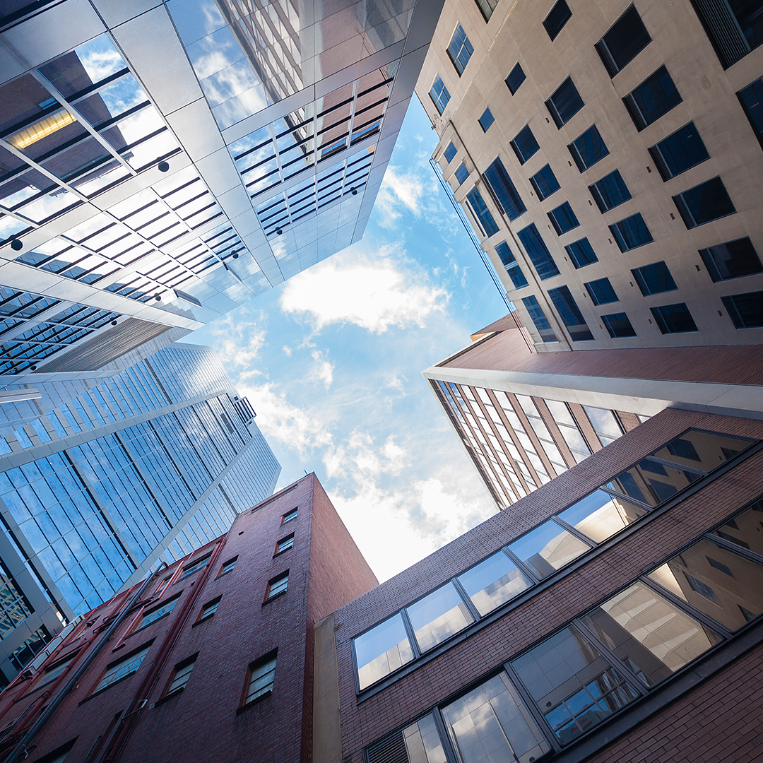 A low-angle shot captures the upward view of various modern and older buildings, with a blue sky and white clouds visible in the centre, giving a sense of being surrounded by the city's architecture.