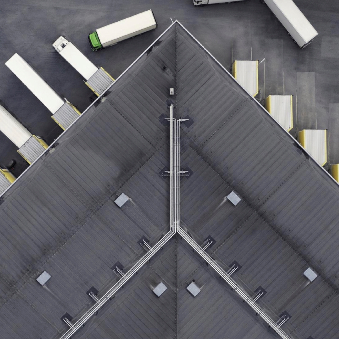 An aerial view captures the sharp, dark grey roof of a warehouse with white piping, surrounded by several semi-trailer trucks, some backed into loading docks and others parked nearby.