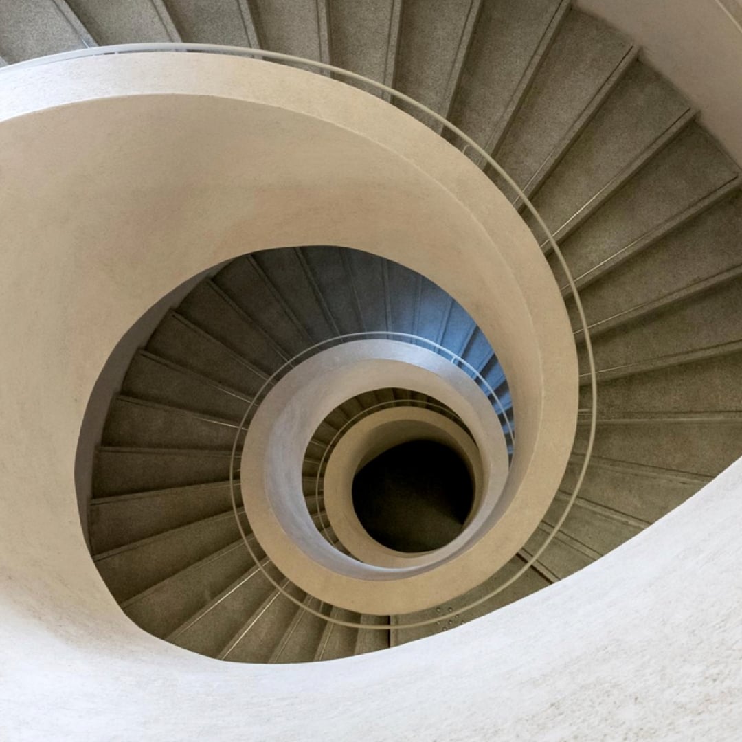 Looking down from above, a striking spiral staircase with grey steps and cream-coloured railings descends into a dark well.