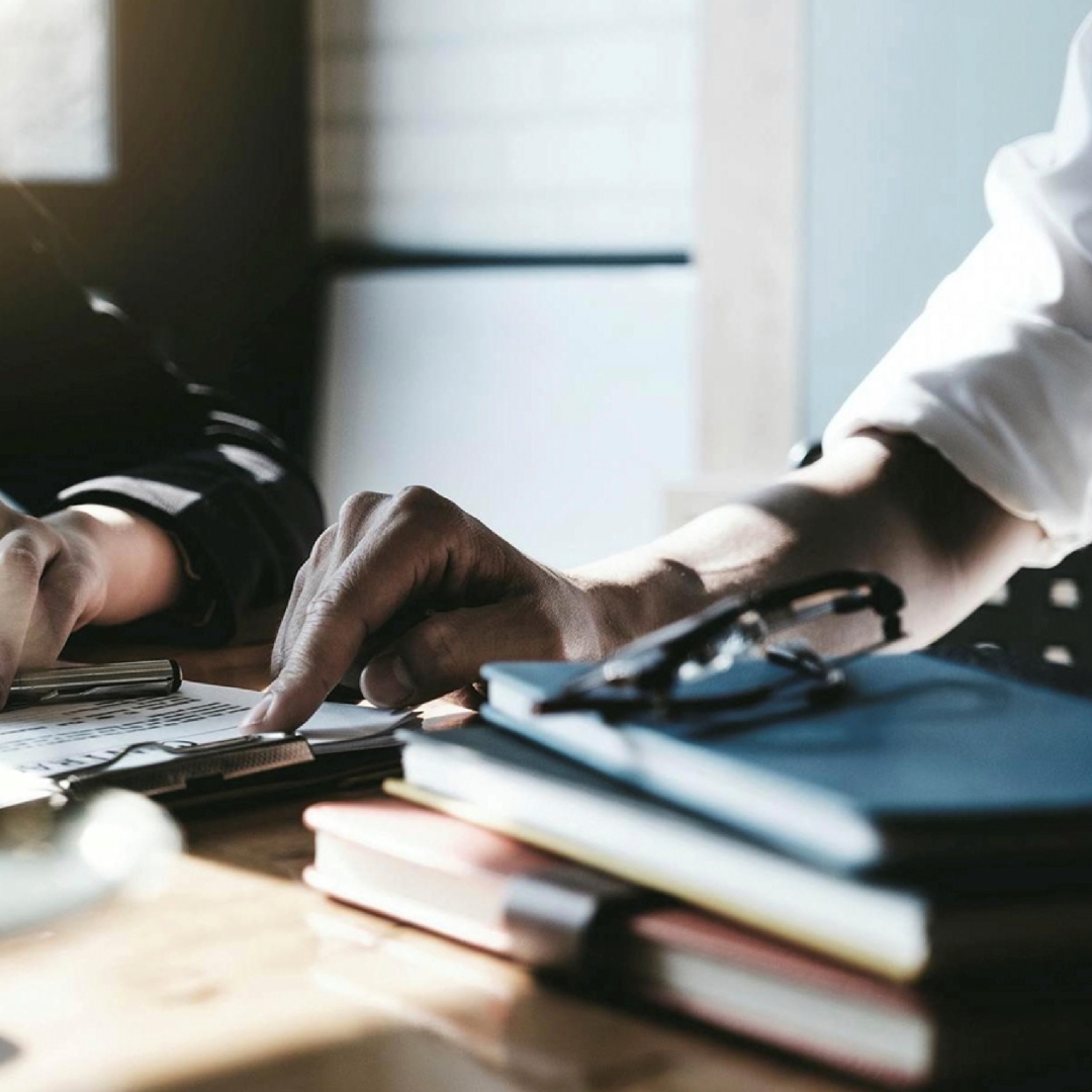 Two individuals are reviewing documents on a wooden desk, with one hand pointing to the text while books and glasses rest nearby.