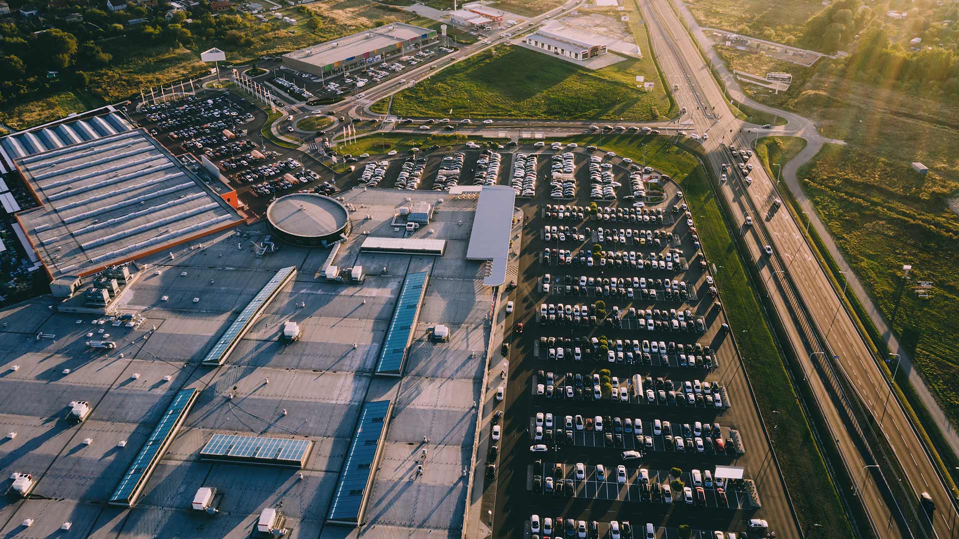 Aerial view of large retail complex with extensive parking filled with cars next to a highway and residential area, including warehouses and retail buildings in sunlight.