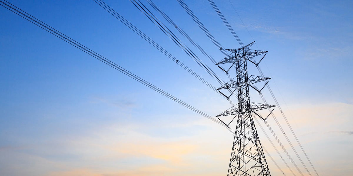 High-voltage transmission tower and overhead power lines against a blue sky, representing electrical grid infrastructure.
