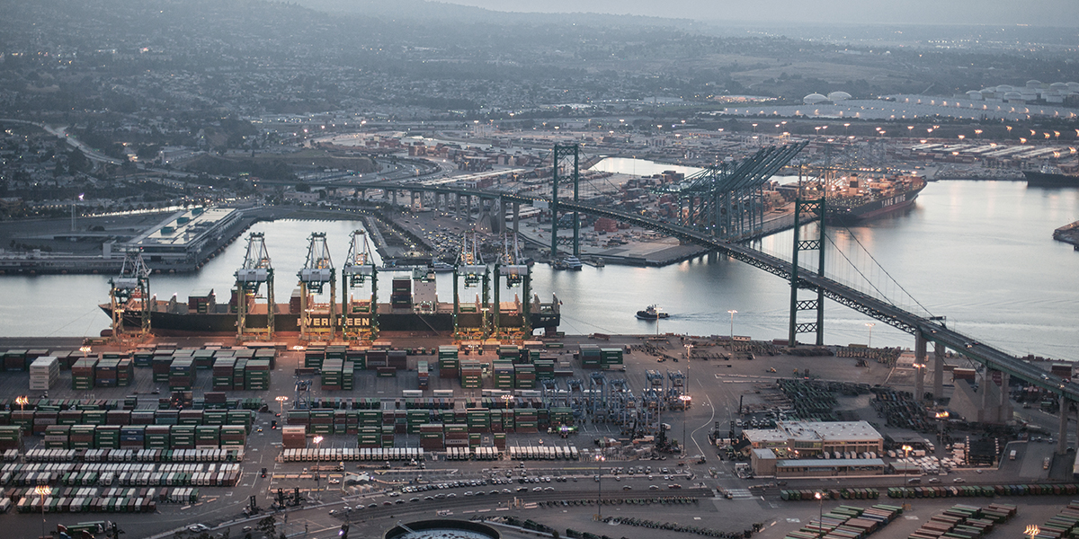 Aerial dusk view of the Port of Los Angeles container terminal with Evergreen cargo ship, ship-to-shore cranes, stacked containers, and Vincent Thomas Bridge.