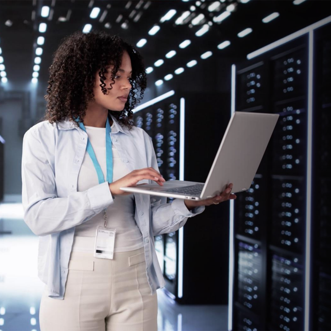 A female IT professional with curly hair types on a laptop in a modern data center, surrounded by glowing server racks.