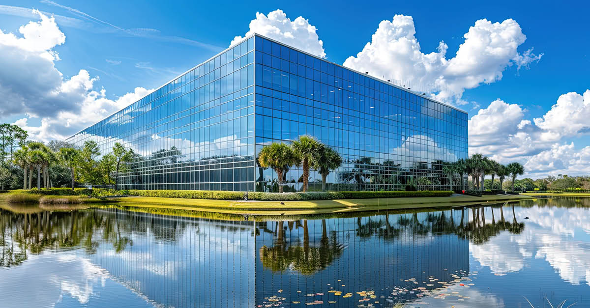 Modern glass office building reflected in a pond, surrounded by landscaping.