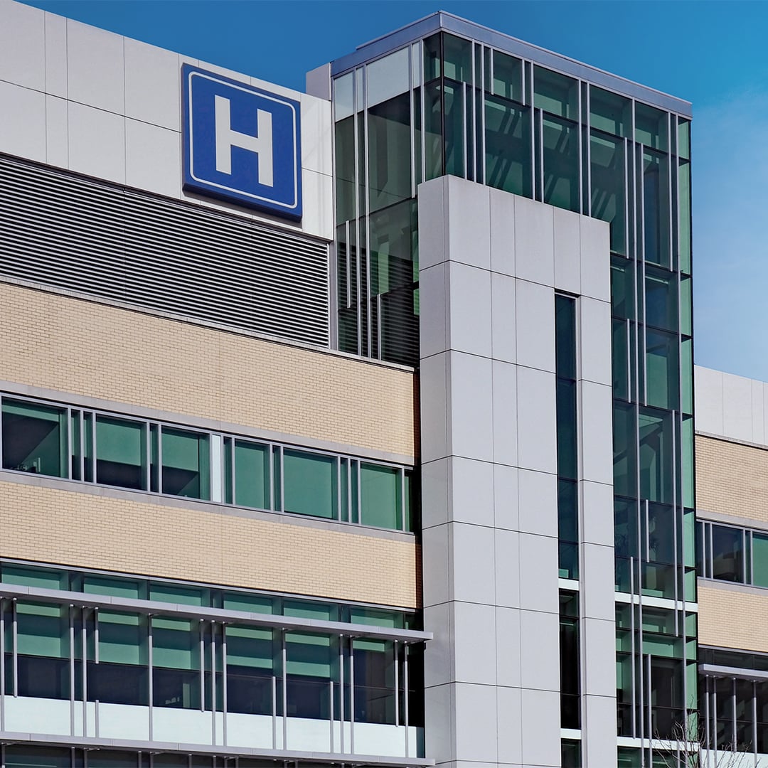 Exterior view of a modern hospital building with glass and metal facade and a blue helipad “H” sign mounted on the upper level.
