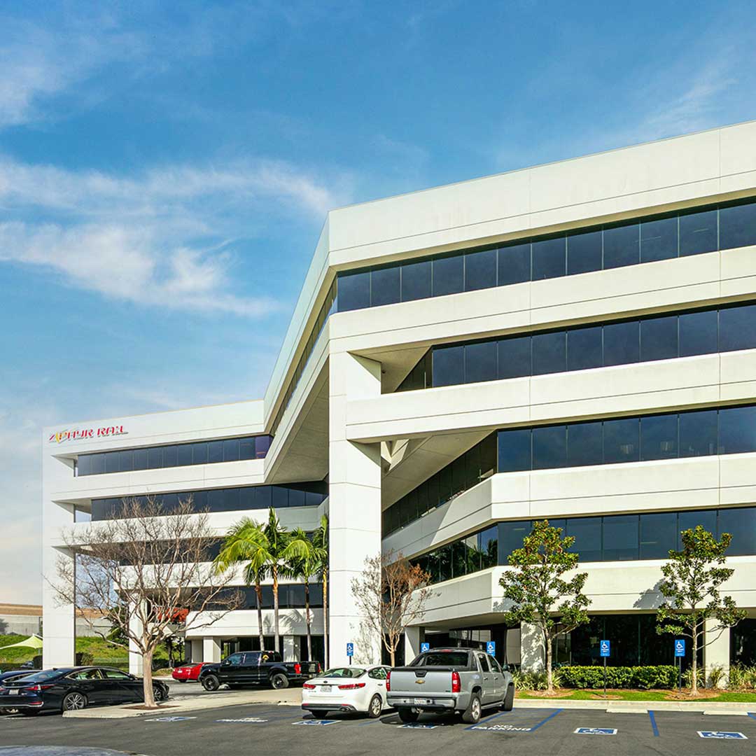 A modern, multi-story office building with large windows and white exterior. Several cars are parked in the parking lot in front of the building. The sky is clear and blue.