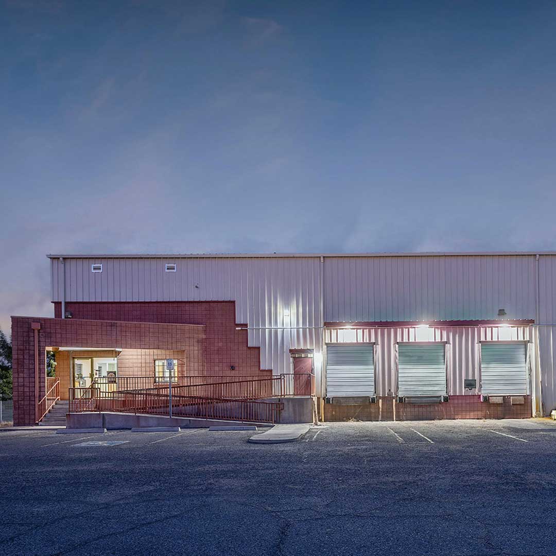 A warehouse building with a combination of brick and metal exterior, featuring three loading dock doors and an entrance with a ramp on the left side. The parking lot in front is empty, and the sky above is clear