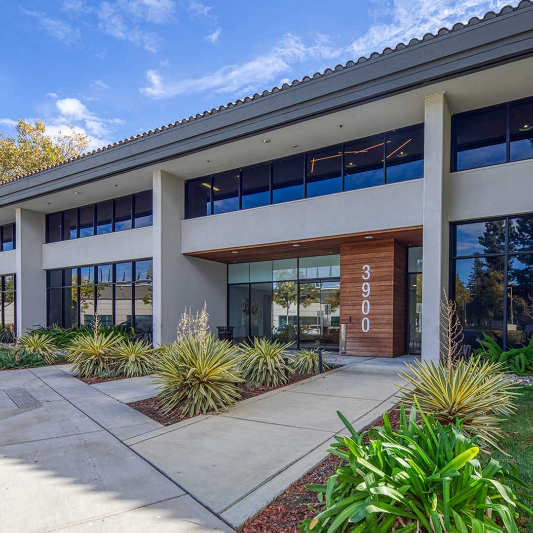 Exterior of a modern commercial building with large glass windows, a flat roof, and a centered entrance. The number '3900' is displayed vertically on a wooden panel beside the door. Landscaped areas with plants and shrubs are in front, under a clear blue sky.