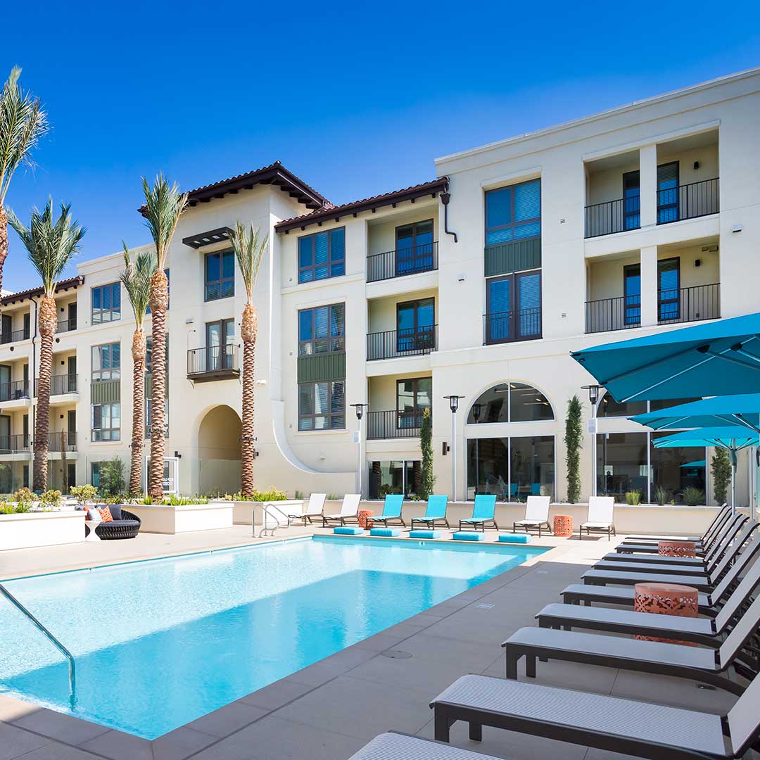 Modern three-story apartment complex with balconies, a swimming pool in the foreground surrounded by lounge chairs, umbrellas, and palm trees, under a clear blue sky.