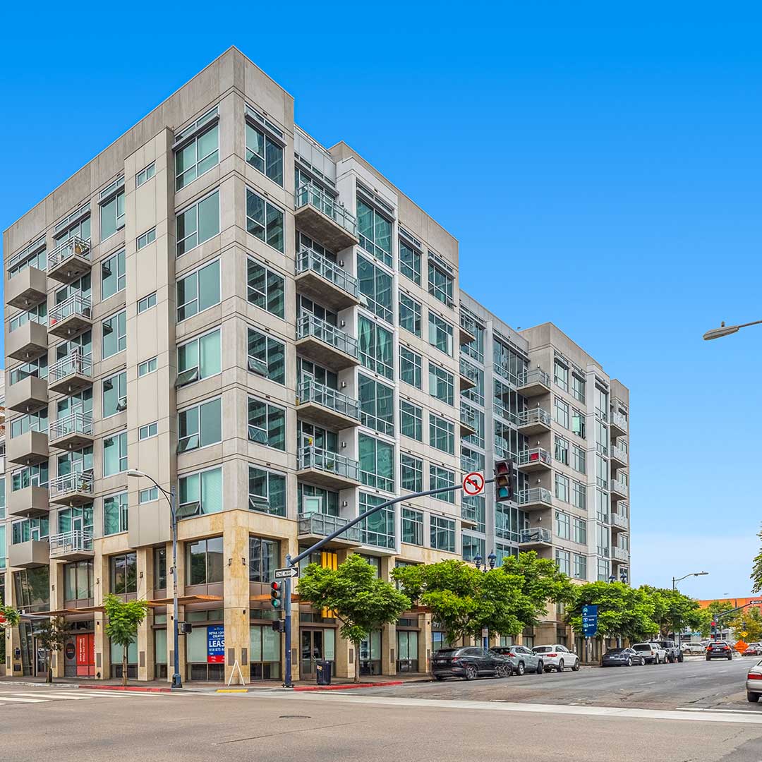 Modern multi-story apartment building with large glass windows and balconies, located at a street corner with trees and parked cars under a clear blue sky.