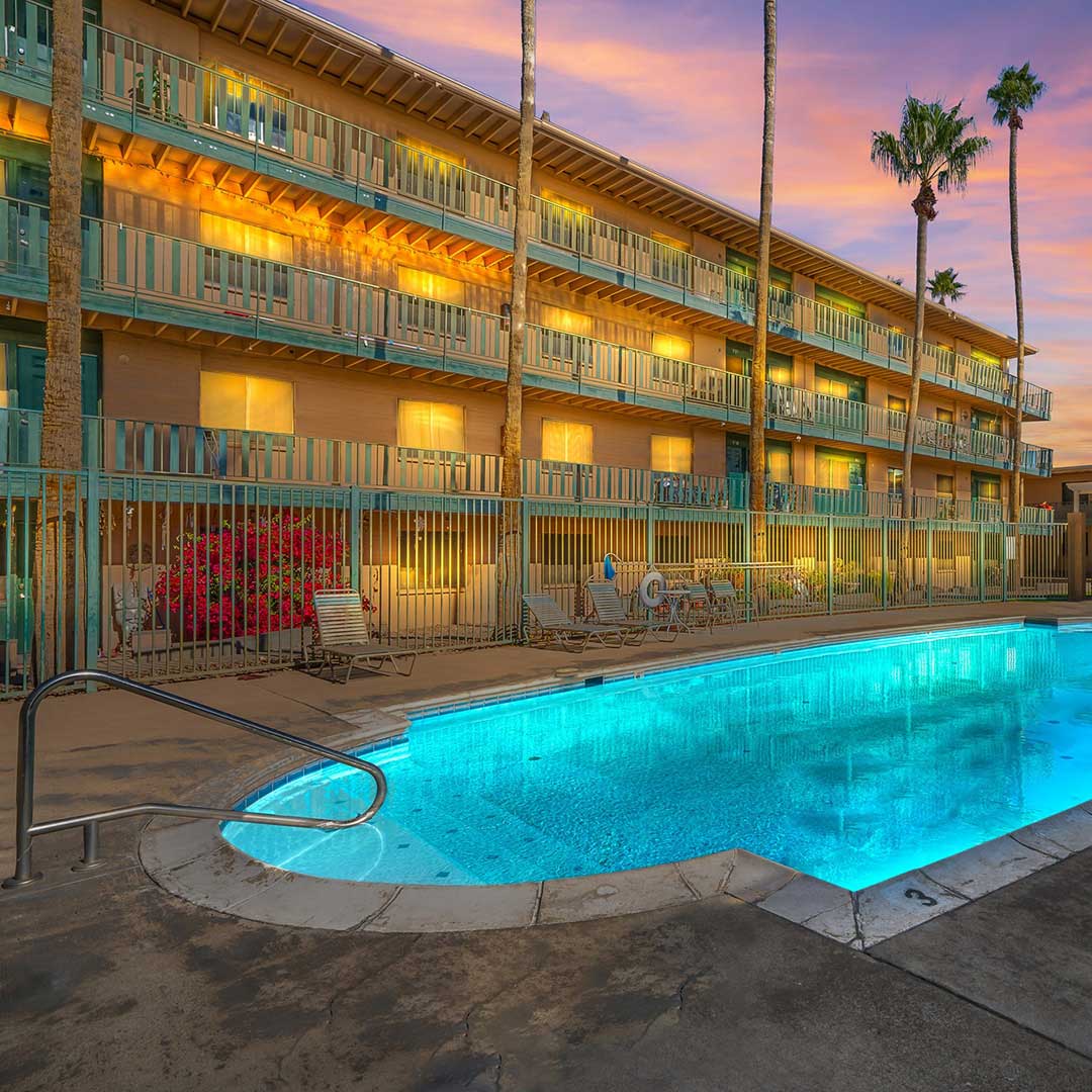 Outdoor swimming pool surrounded by palm trees and lounge chairs, in front of a multi-story building with balconies and warm lighting, under a colorful sunrise or sunset sky.