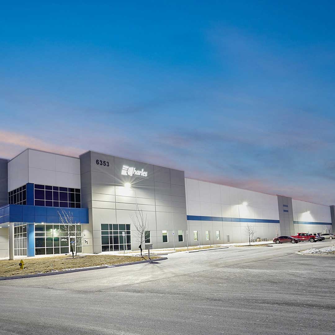 Modern industrial building labeled '6353 Charles' with large windows, exterior lighting, small trees in front, and parked cars to the right, under a clear gradient sky.
