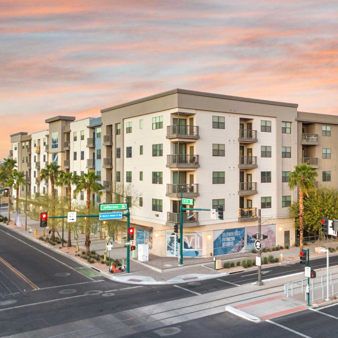 Modern multi-story apartment building at the corner of Jefferson Street and 1st Avenue, with balconies, large windows, palm trees, and a fitness studio banner at street level. Traffic lights and street signs are visible, with a colorful pink and orange sky in the background.