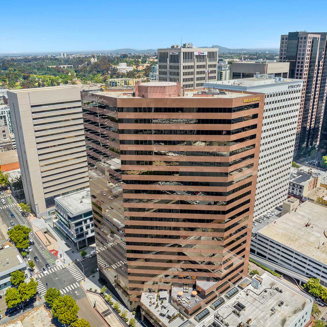 Aerial view of a cityscape featuring a tall brown glass building with the Wells Fargo logo, surrounded by office buildings, streets with traffic and pedestrians, and distant hills under a clear blue sky.