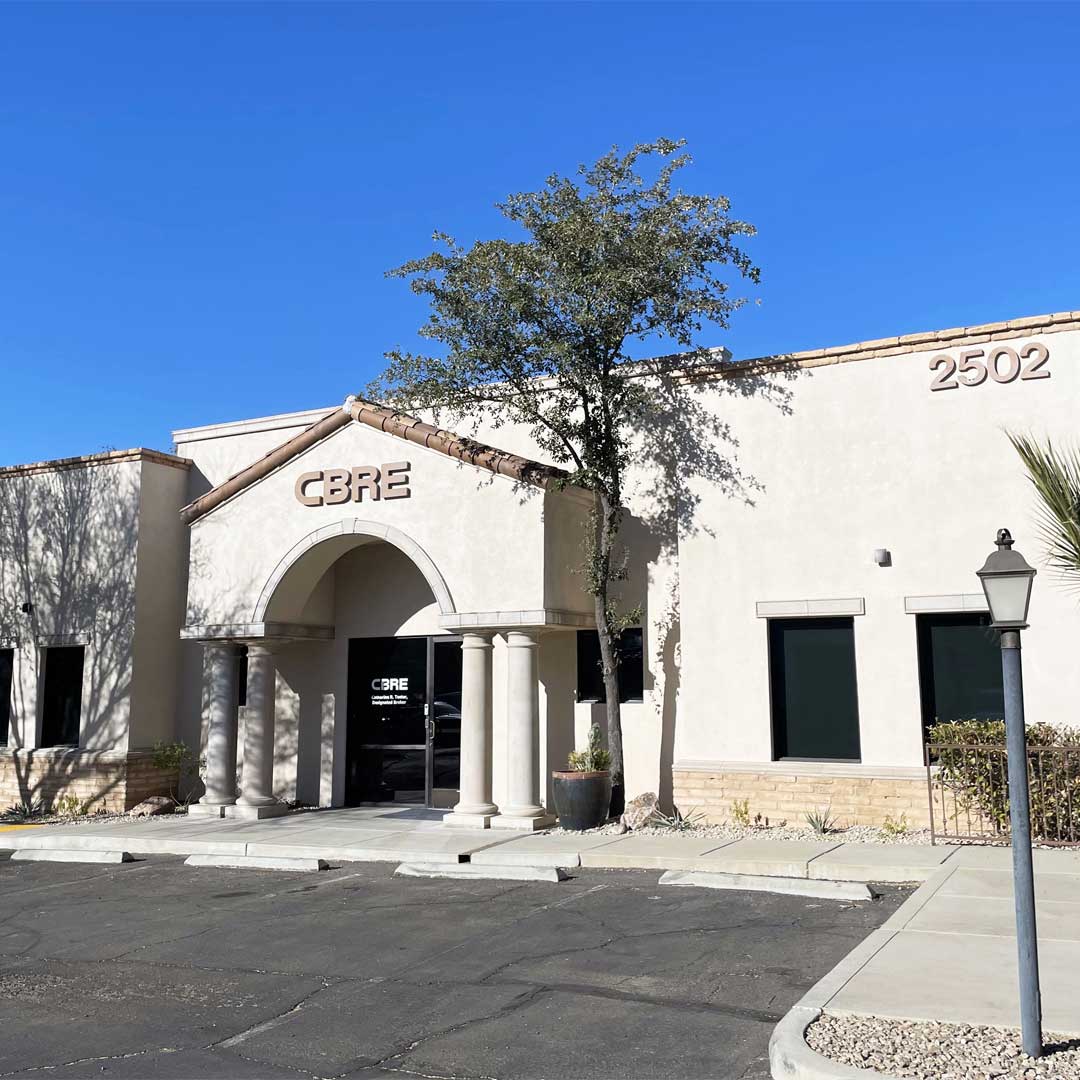 Commercial building with 'CBRE' signage above an arched entrance, beige exterior, and the number '2502' on the right side. Surrounded by trees, plants, and a lamp post under a clear blue sky.