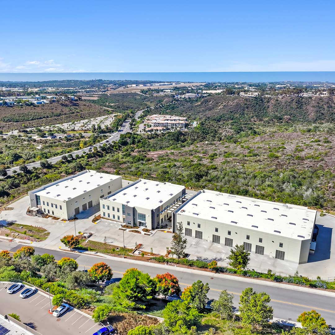Aerial view of three large industrial buildings surrounded by greenery and hills under a clear blue sky.