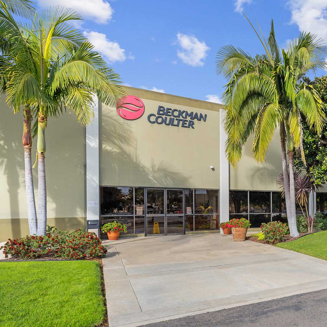 Beckman Coulter building entrance with glass doors, palm trees, and potted plants under a blue sky.