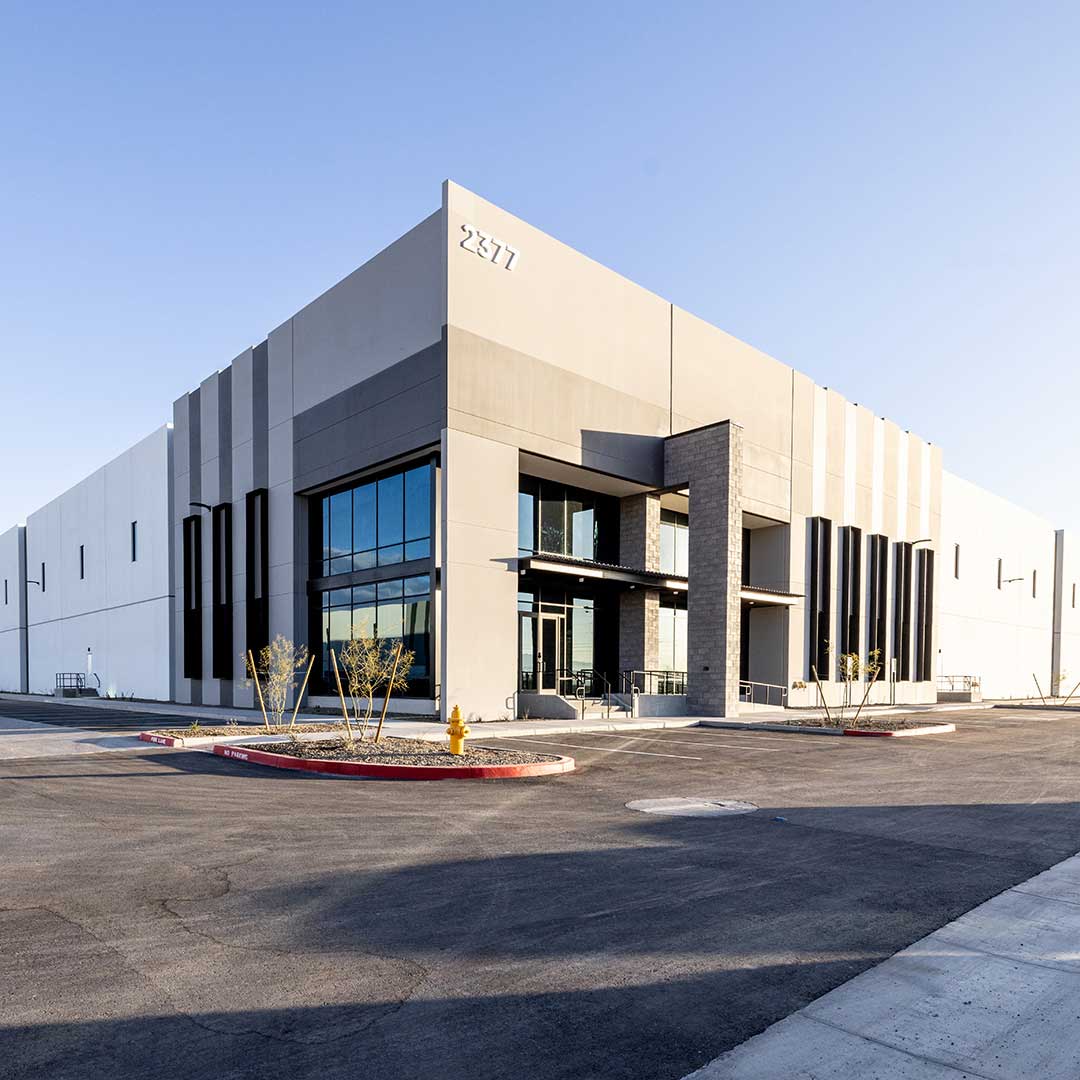 Modern commercial building with large glass windows, vertical lines, and the number '2377' displayed on the upper corner. Landscaped entrance with plants and a fire hydrant in front, surrounded by a paved area.