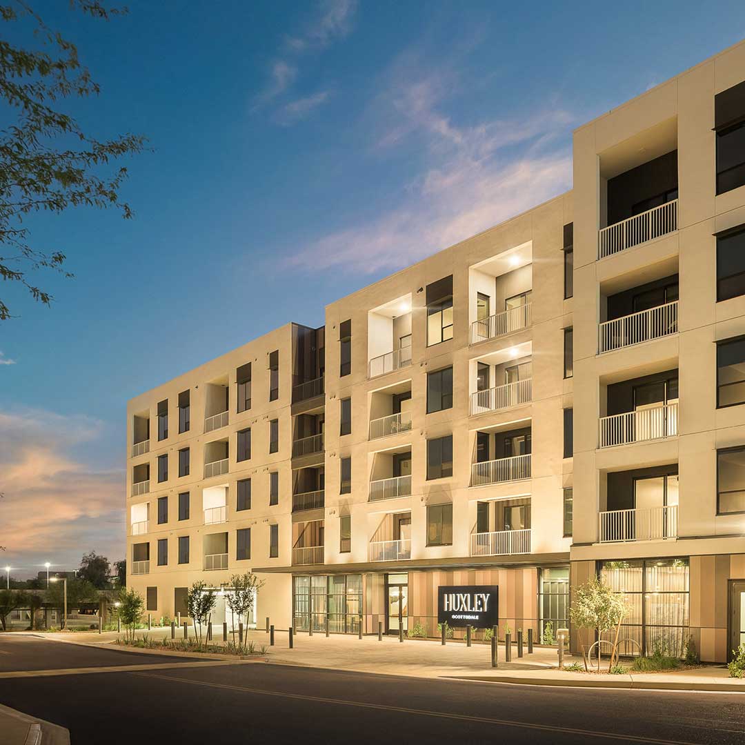 Modern multi-story apartment building named 'HUXLEY' with balconies and large windows, illuminated during sunset or sunrise. The sky has a blue-orange gradient, and the surrounding area includes trees, plants, and an empty street.