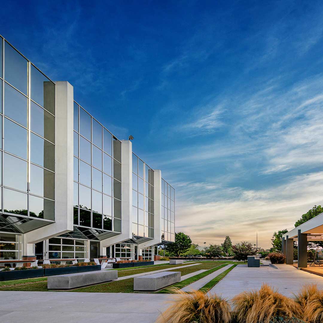 Modern building with large glass windows reflecting a blue sky, vertical concrete columns, and a landscaped area with grass, trees, and benches under a clear sky with wispy clouds.