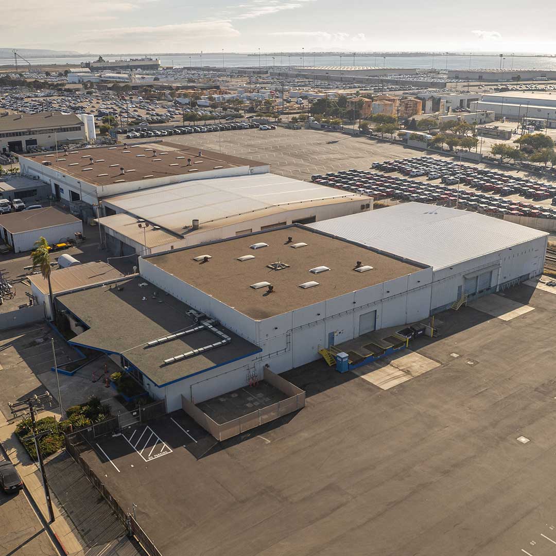 Aerial view of an industrial area with multiple large warehouse buildings, parking lots filled with cars, and rooftop equipment on the foreground building. Clear sky with some clouds on the horizon.