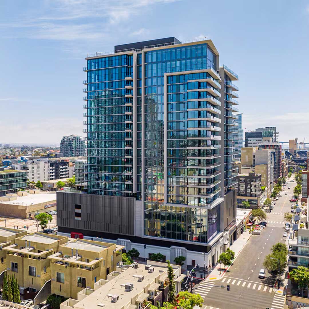 Modern high-rise building with a glass facade in an urban area, featuring multiple floors and balconies, surrounded by other buildings and streets under a clear daytime sky.