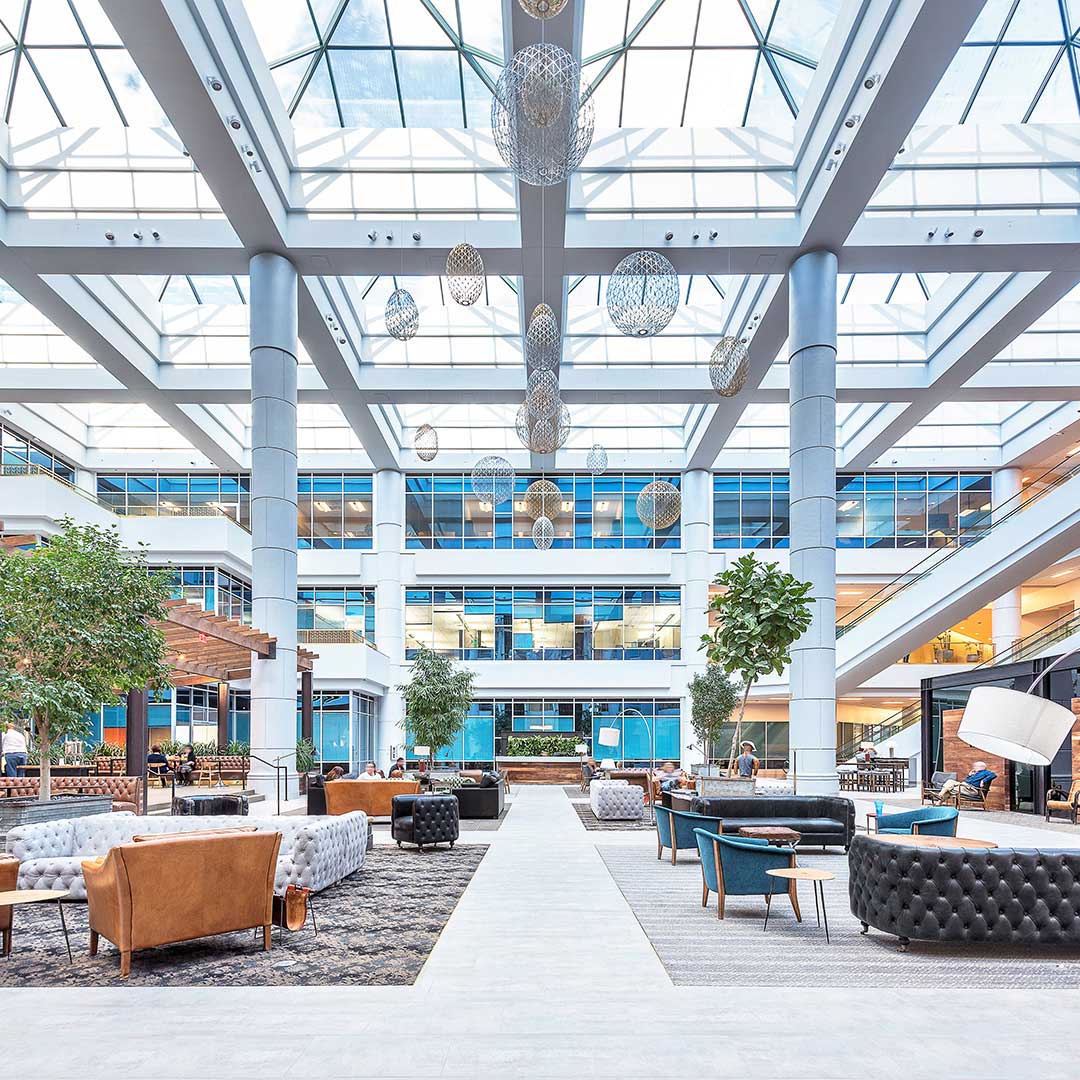 Spacious modern indoor atrium with a high glass ceiling, large windows, various seating arrangements, potted trees, and spherical hanging light fixtures, likely part of a multi-story office or commercial building.