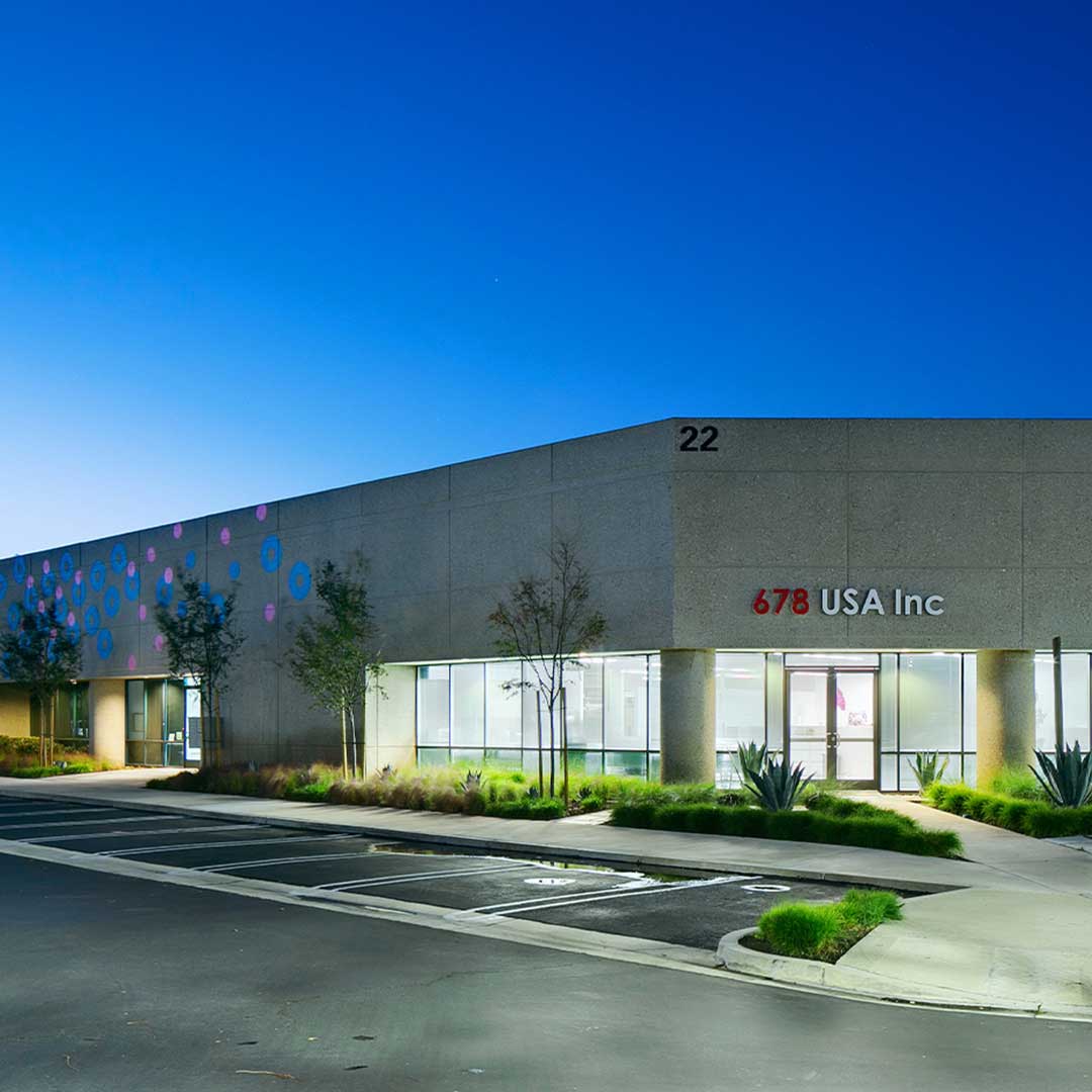 Modern commercial building with address number 22 and signage reading '678 USA Inc' on the front. The building has large windows, landscaped greenery, and a parking lot in front under a clear sky.
