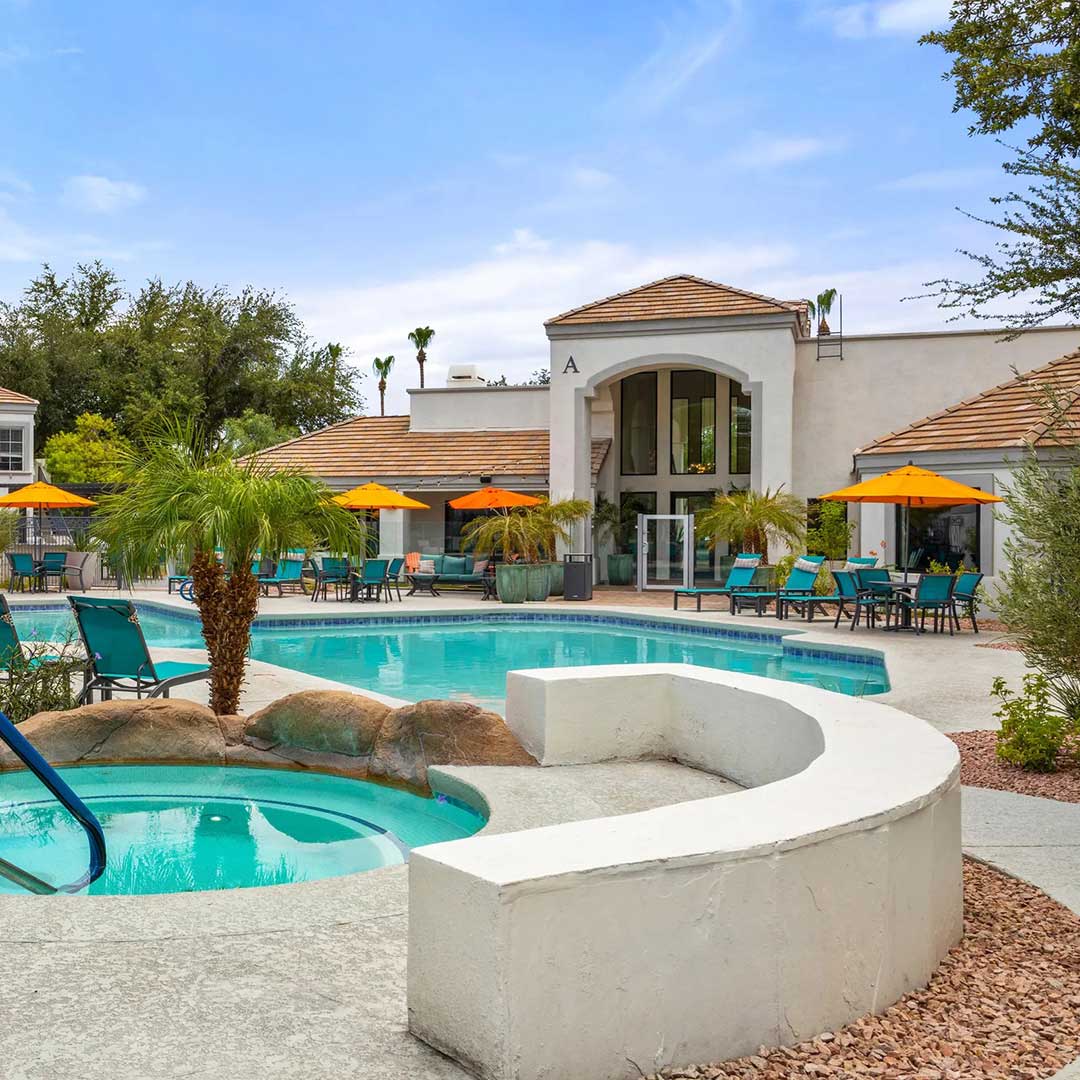 Outdoor pool area with a main swimming pool and a smaller circular hot tub, surrounded by lounge chairs and orange umbrellas. A large white building with terracotta roof tiles and an entrance labeled 'A' is in the background, with palm trees and greenery adding to the tropical setting under a partly cloudy sky.