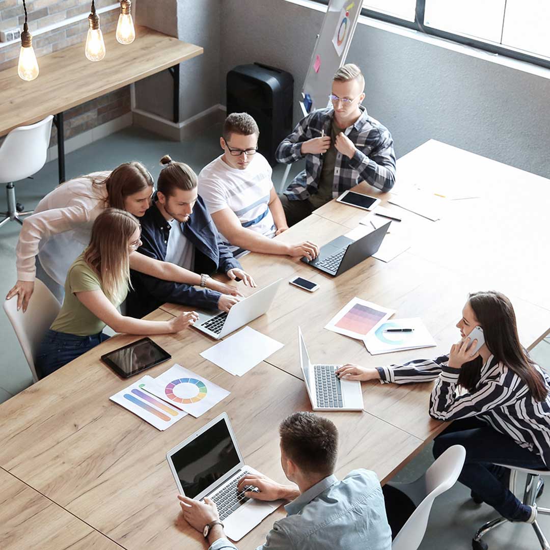 Group of people collaborating in a modern office, seated around a large wooden table with laptops, tablets, and documents, under hanging lights and near large windows.
