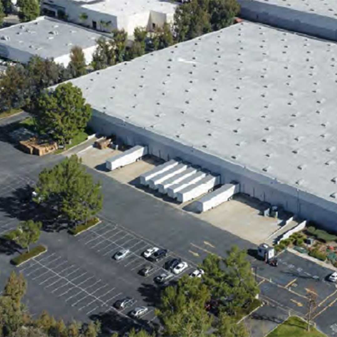 Aerial view of a large industrial warehouse with a white roof, multiple loading docks with white trailers, and a surrounding parking lot with scattered cars and trees.
