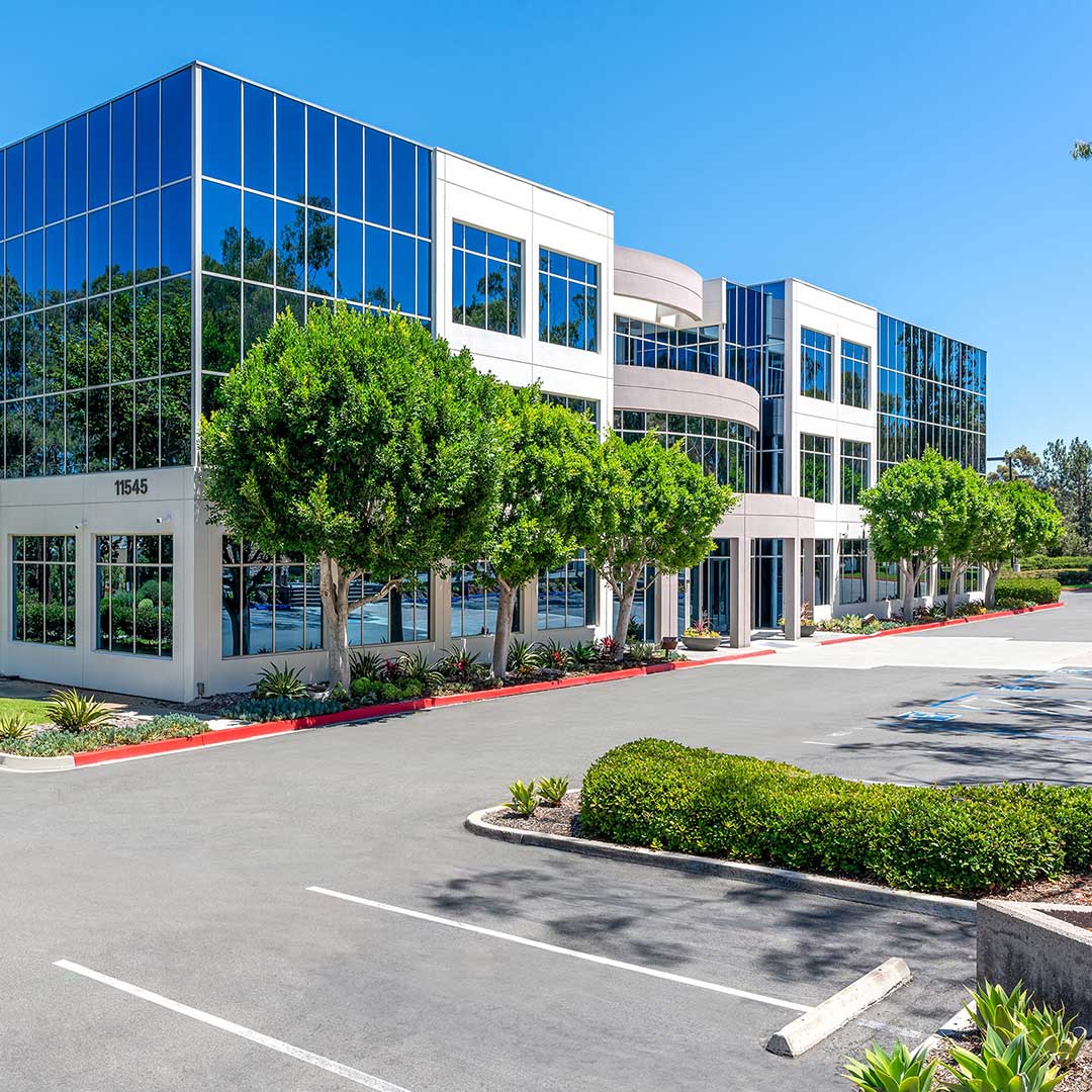 Three-story modern office building with large glass windows reflecting trees and sky, surrounded by landscaped greenery and a parking lot. The address number '11545' is visible on the building.