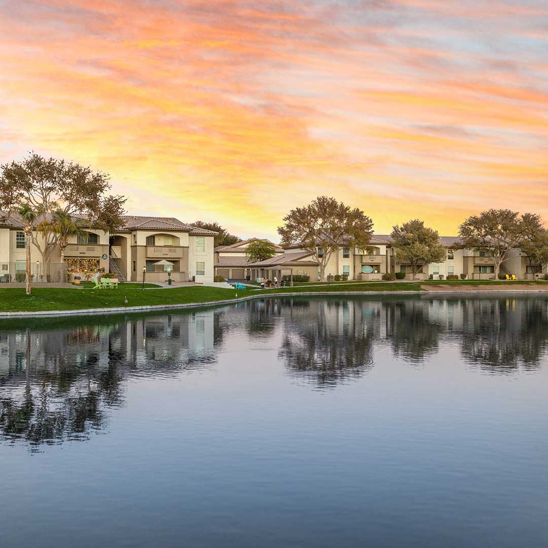 Serene lakeside view at sunrise or sunset with residential buildings reflected in calm water, surrounded by trees and a colorful sky in shades of orange, pink, and yellow.